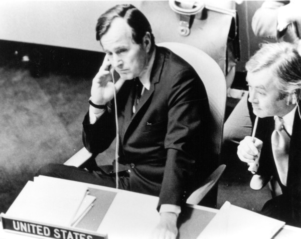 Photographic print of George Bush seated at a United Nations delegation table marked 'United States,' holding a headset to his ear, with a colleague beside him