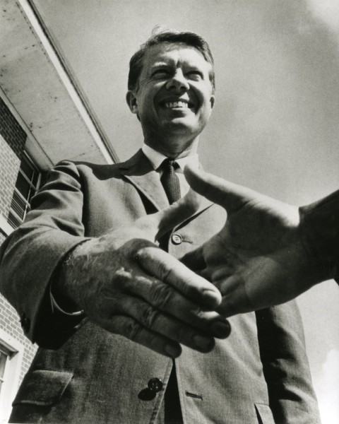 Photographic print of Jimmy Carter in a suit and tie, smiling broadly while shaking hands, shot from a low angle against an overcast sky