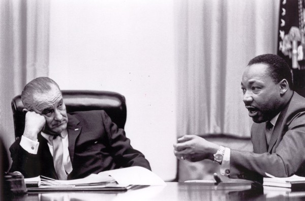 Photographic print of Dr. Martin Luther King, Jr. speaking and gesturing across a table while President Lyndon B. Johnson listens, chin resting on his hand