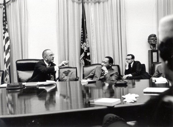 Photographic print of President Johnson gesturing while speaking at a large conference table, with Martin Luther King, Jr. and other attendees seated nearby. An American flag and a Presidential flag hang behind them, with a bust on a pedestal in the background