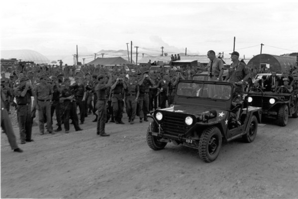 Photographic print of President Johnson and General Westmoreland standing in a military jeep, waving to a large crowd of soldiers at a base in Vietnam