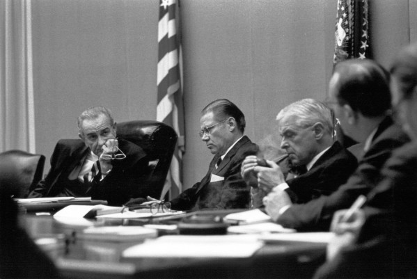 Photographic print of President Lyndon B. Johnson seated at a conference table with Robert McNamara, Paul Nitze, and General Earle Wheeler, American flags visible behind them