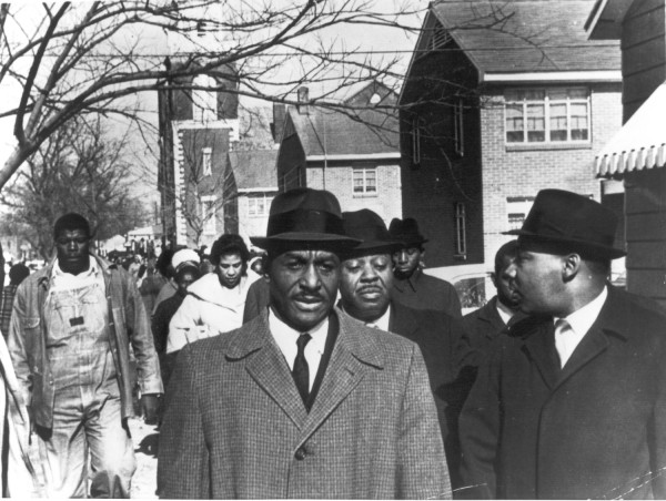 Photographic print of Rev. Fred Shuttlesworth, Martin Luther King Jr., and Ralph Abernathy leading a crowd of marchers along a street lined with brick buildings
