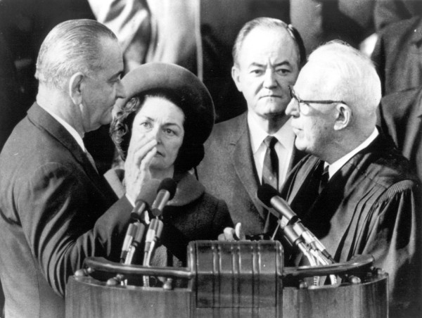 Photographic print of Lyndon B. Johnson facing Chief Justice Earl Warren in judicial robes at a podium with microphones. Lady Bird Johnson and Vice President Hubert Humphrey look on behind them