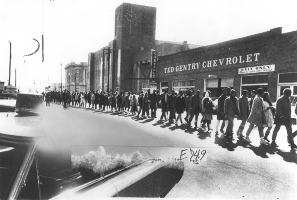 Photographic print of a long column of young people marching along a street past a brick building marked Ted Gentry Chevrolet, Selma, Alabama