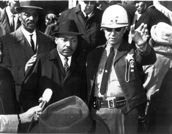 Photographic print of Dr. Martin Luther King, Jr. in a hat and overcoat facing a uniformed deputy sheriff with a star badge, who gestures with one hand. A crowd and microphone are visible