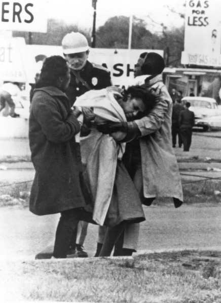 Photographic print showing two people supporting an unconscious woman on a roadside as a uniformed state trooper in a white helmet stands behind them