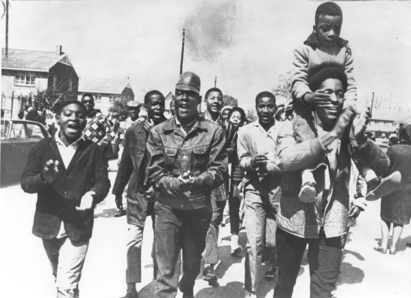 Photographic print of a group of young marchers walking, singing, and clapping on a street in Selma, Alabama. One child rides on an adult's shoulders as buildings and utility poles line the background