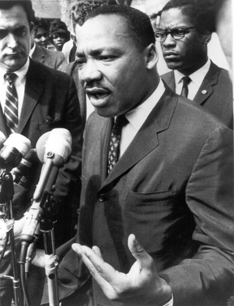 Photographic print of Dr. Martin Luther King, Jr. speaking into multiple microphones outdoors, gesturing with his hands, surrounded by several people in suits