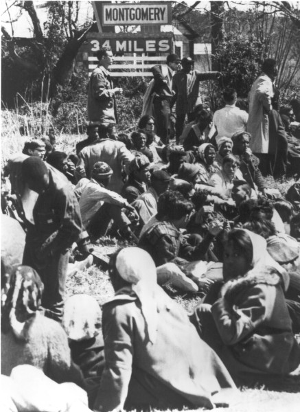 Photographic print of a large group of civil rights marchers sitting and resting on a roadside embankment. A sign reading "Montgomery 34 Miles" is visible behind them