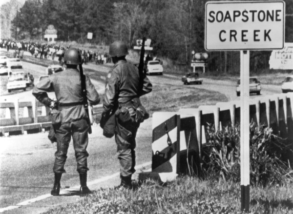 Photographic print of two armed, helmeted soldiers standing on a bridge beside a Soapstone Creek sign, as a large crowd of marchers advances along the road in the background