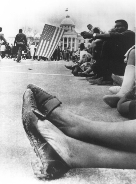 Photographic print showing worn, muddy shoes in the foreground, marchers resting along a street, an American flag, and the Alabama state capitol dome in the background