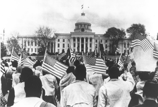 Photographic print of a large crowd waving American flags before the domed Alabama state capitol building. Some marchers hold signs, including one reading "Freedom Now"