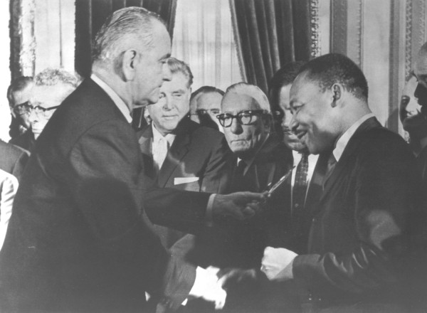 Photographic print of President Johnson handing a pen to Dr. Martin Luther King, with Rep. Claude Pepper wearing glasses and Rev. Ralph Abernathy visible among onlookers