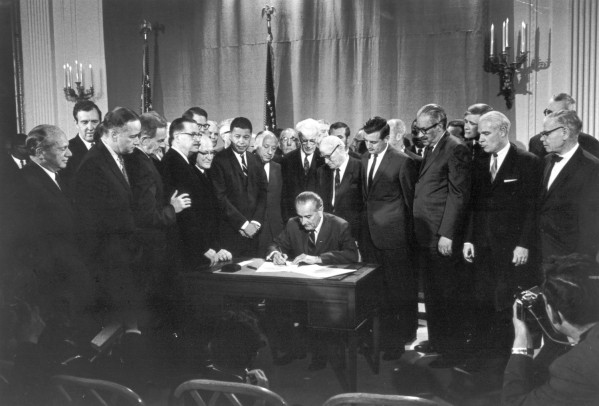 Photographic print of President Lyndon B. Johnson seated at a desk signing a document, surrounded by a large crowd of suited politicians in a formal room