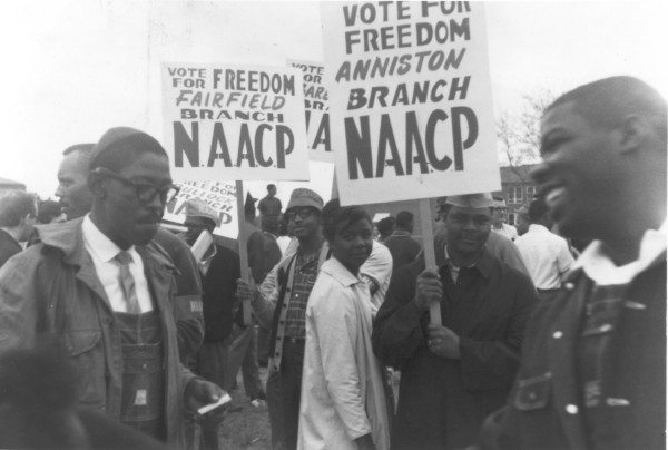 Photographic print of a crowd of NAACP supporters marching outdoors, carrying signs reading "Vote for Freedom" from the Fairfield and Anniston branches of the NAACP
