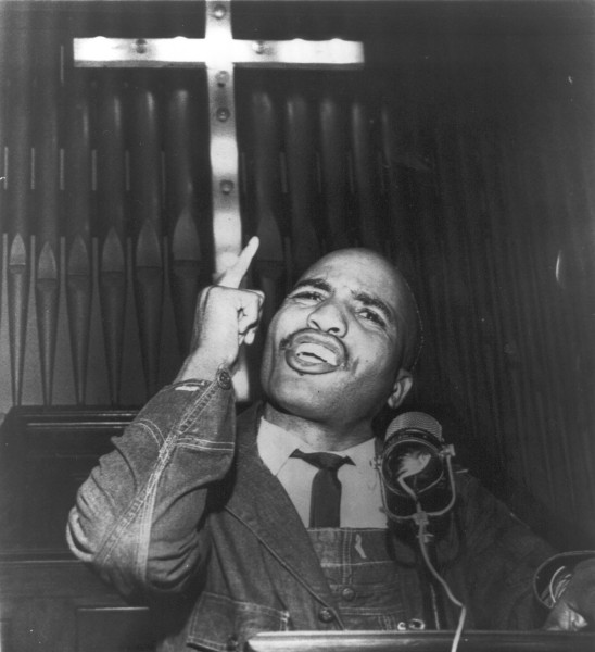 Photographic print of James Bevel speaking at a church podium with a microphone, one finger raised upward toward a cross on the wall behind him