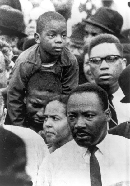 Photographic print of a young boy perched on a man's shoulders amid a dense crowd of marchers. Dr. Martin Luther King, Jr. is visible in the foreground wearing a white shirt and tie
