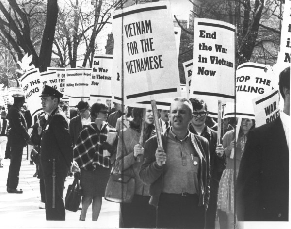 Photographic print of protesters marching on a sidewalk carrying signs reading "Vietnam for the Vietnamese," "End the War in Vietnam Now," and "Stop the Killing." A uniformed officer stands at left as bare trees line the background