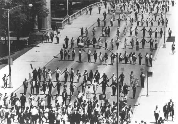 Photographic print of hundreds of marchers holding hands in long rows spanning a wide urban street, with protest signs visible in the foreground