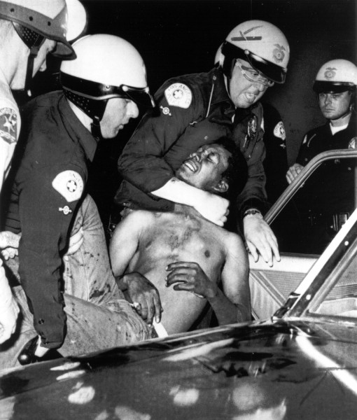 Photographic print showing several Los Angeles police officers in helmets forcibly placing a shirtless man into a vehicle at night