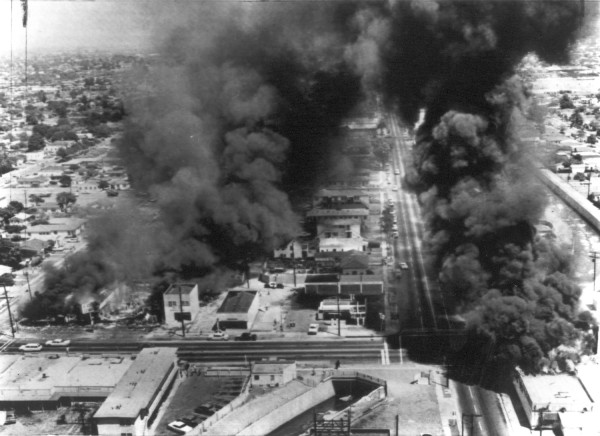 Photographic print aerial view of three buildings engulfed in flames, sending large columns of dark smoke into the sky over a dense urban neighborhood