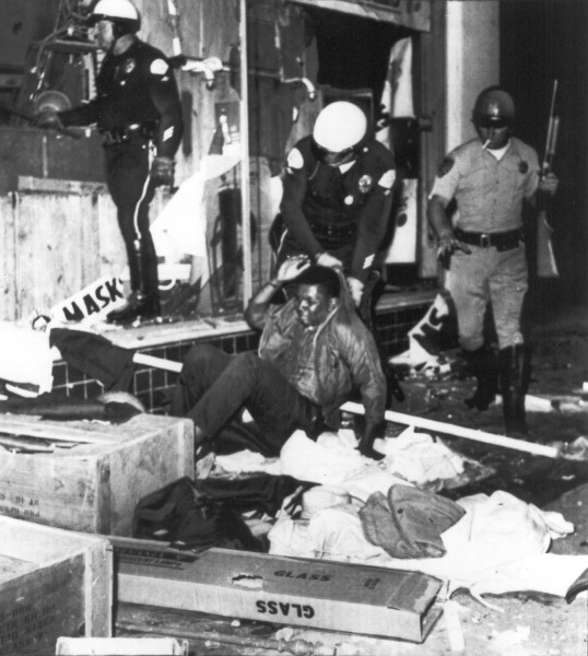 Photographic print of helmeted police officers dragging a man on the ground outside a storefront strewn with scattered debris and overturned boxes