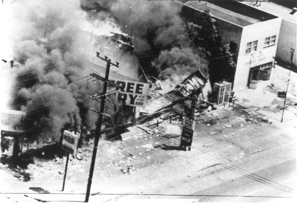 Photographic print, aerial view of three stores collapsed and engulfed in flames on a city block, with large plumes of dark smoke rising above the rubble