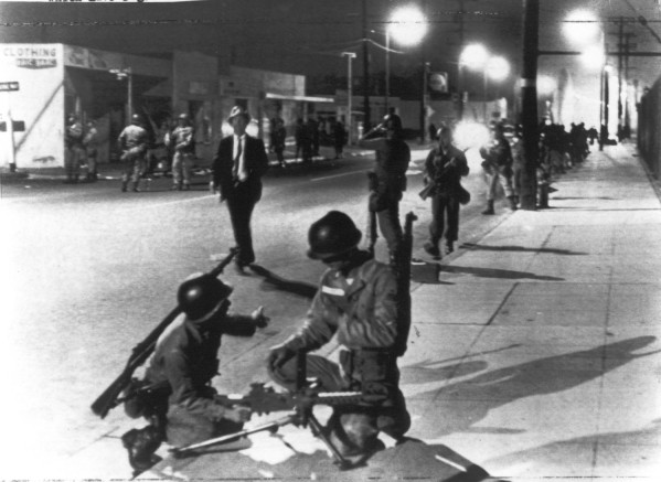 Photographic print of California National Guardsmen in helmets and fatigues crouching and standing along a night-lit urban street, rifles visible