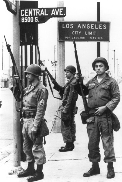 Photographic print of three uniformed, helmeted National Guardsmen holding rifles on a sidewalk beneath a Los Angeles City Limit sign at Central Ave. and 8500 S.