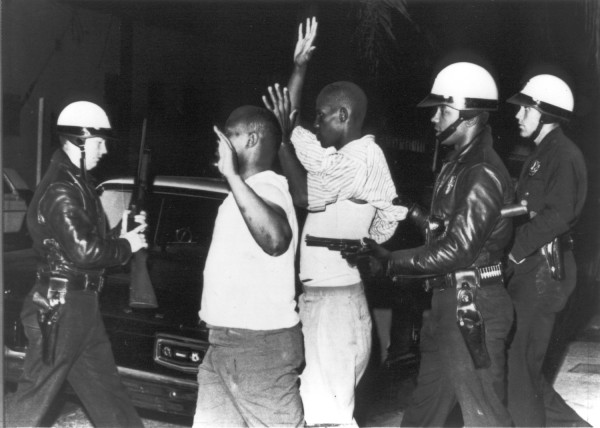 Photographic print of three helmeted officers holding guns on two men with hands raised, outdoors at night beside a parked car