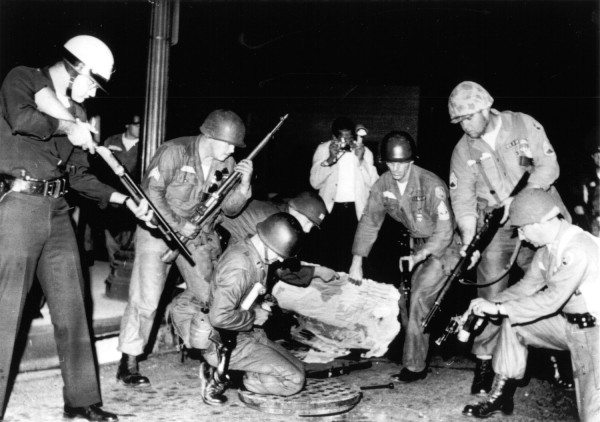 Photographic print of armed police officers and helmeted National Guardsmen surrounding an open street manhole at night, rifles raised