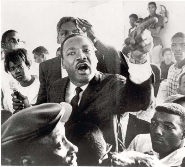 Photographic print of Martin Luther King, Jr. speaking passionately, arm outstretched and pointing, surrounded by a crowd of onlookers in Watts, Los Angeles