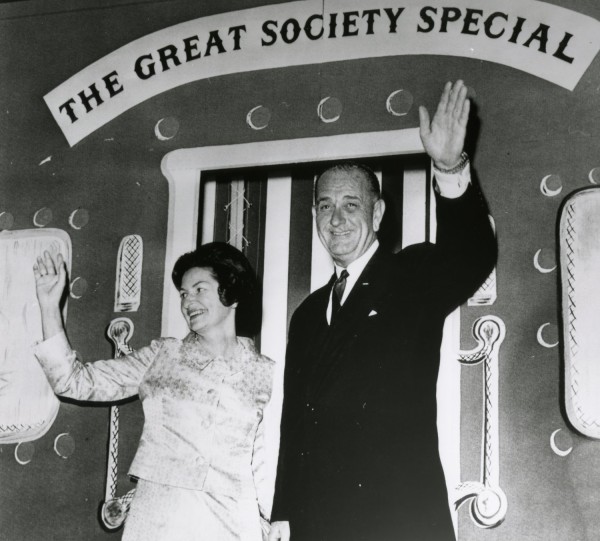Photographic print of President Johnson and Lady Bird Johnson holding hands and waving beneath a sign reading "The Great Society Special"