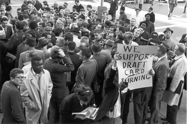 Photographic print of a dense outdoor crowd, with a man holding a half-torn sign reading "We Support Draft Card Burning" visible on the right