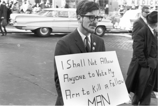 Photographic print of a young bearded man in a suit and tie holding a sign reading "I Shall Not Allow Anyone to Vote My Arm to Kill a Fellow MAN" on a city street