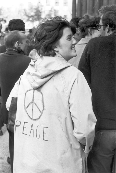 Photographic print of a woman in a crowd wearing a light jacket with a hand-drawn peace symbol and the word "PEACE" on the back