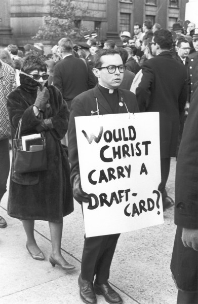 Photographic print of a priest in clerical collar holding a sign reading "Would Christ Carry a Draft-Card?" amid a crowd on a city sidewalk