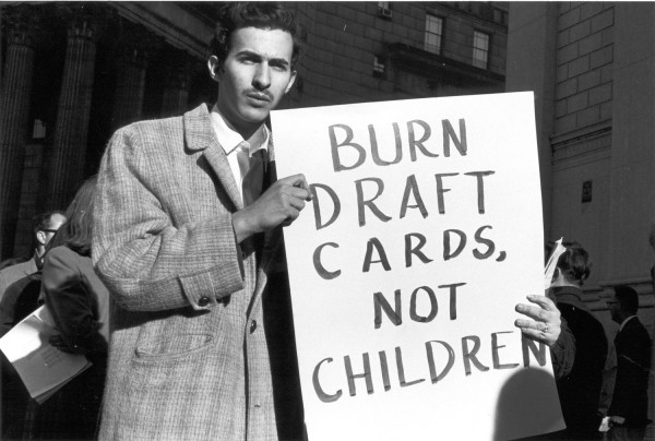 Photographic print of a man in a wool coat holding a large sign reading "Burn Draft Cards, Not Children" outdoors near a columned building