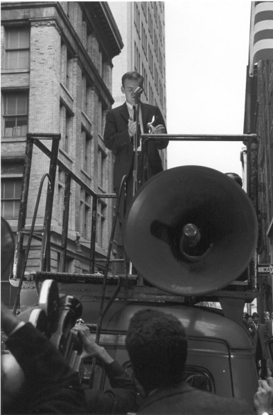 Photographic print of David Miller standing on a raised platform, holding a lighter to a small card at a microphone, with a crowd and city buildings below