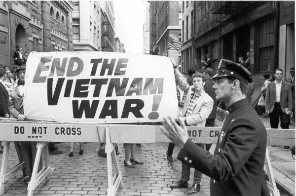 Photographic print of protesters holding a large "End the Vietnam War!" banner behind police barricades on a cobblestone street. A uniformed police officer stands in the foreground near the barriers