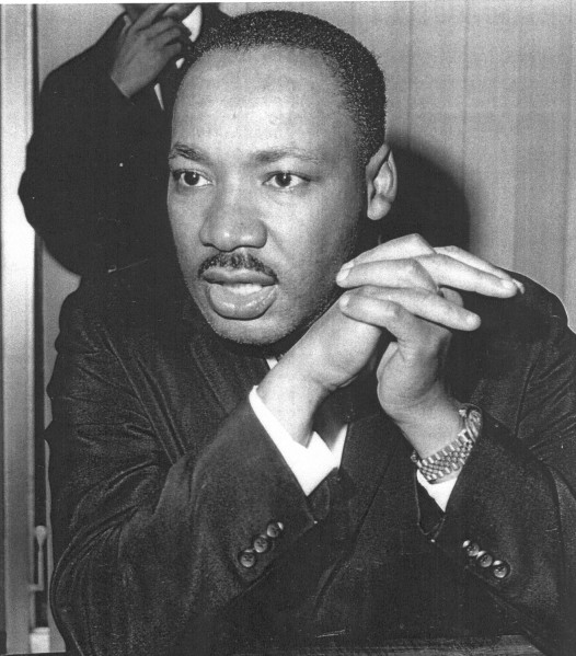 Photographic print of Martin Luther King, Jr. in a dark suit, hands clasped near his chin, speaking at a press gathering. A second figure is partially visible in the background.