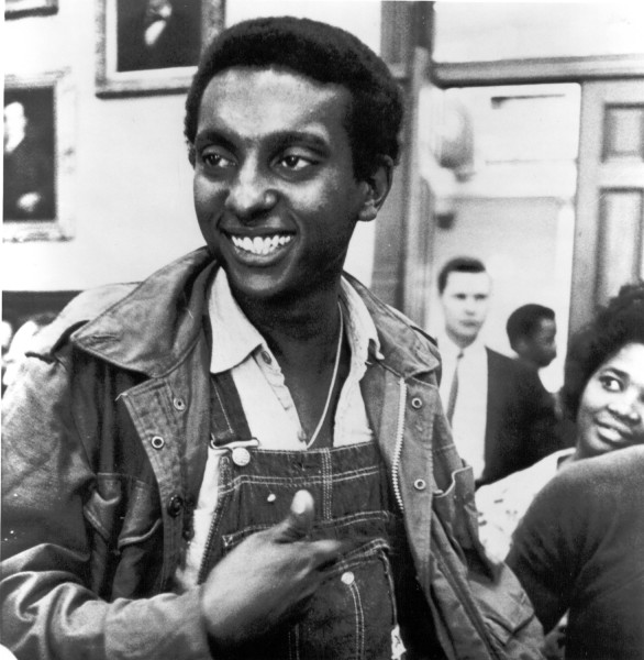 Photographic print of Stokely Carmichael smiling broadly, wearing denim overalls and a jacket, gesturing while speaking in a crowded indoor setting