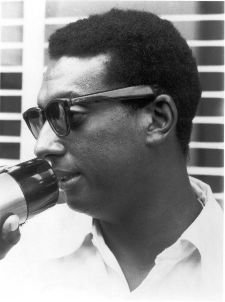Photographic print of Stokely Carmichael in profile, wearing sunglasses and a white shirt, speaking into a bullhorn