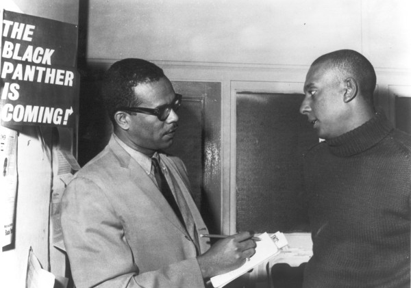 Photographic print of Stanley S. Scott, holding a notepad and pen, speaking with Stokely Carmichael indoors. A poster reading "The Black Panther Is Coming!" is visible on the wall behind them