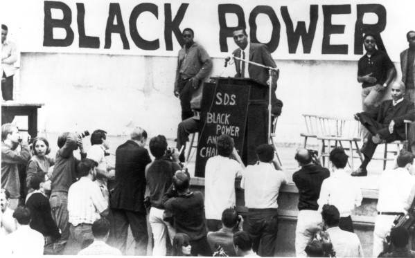 Photographic print of Stokely Carmichael speaking at a podium labeled 'S.D.S. Black Power' beneath a large 'BLACK POWER' banner. A crowd of photographers and attendees surrounds the outdoor stage