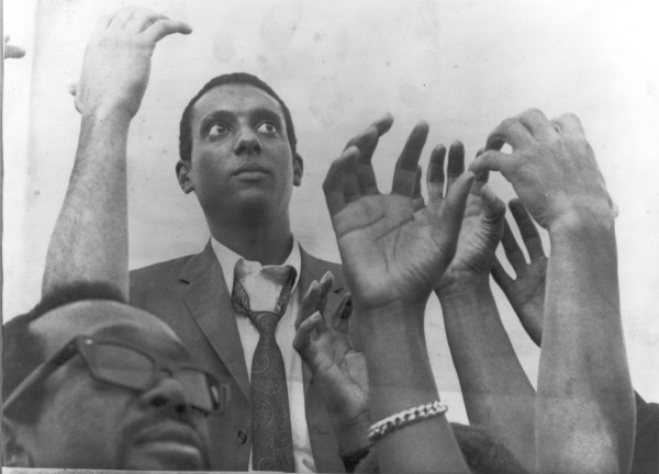 Photographic print of Stokely Carmichael in a suit and tie, looking upward, surrounded by raised and outstretched hands of rally attendees