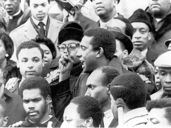Photographic print of Stokely Carmichael speaking in a dense outdoor crowd, one hand raised with a pointed finger, mouth open mid-speech