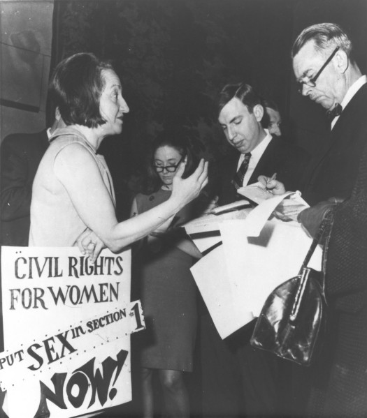 Photographic print of Betty Friedan speaking to reporters taking notes, beside a sign reading "Civil Rights for Women — Put Sex in Section #1 Now!"