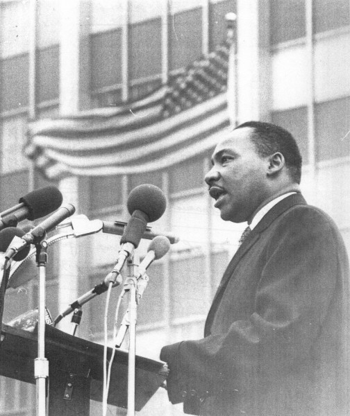Photographic print of Martin Luther King, Jr. speaking at a podium with multiple microphones. An American flag waves in the background behind him.
